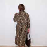 Woman wearing a patterned dress holding a handbag against a white wall.