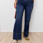 Person wearing blue jeans with a gold belt and brown shoes on a light wooden floor.