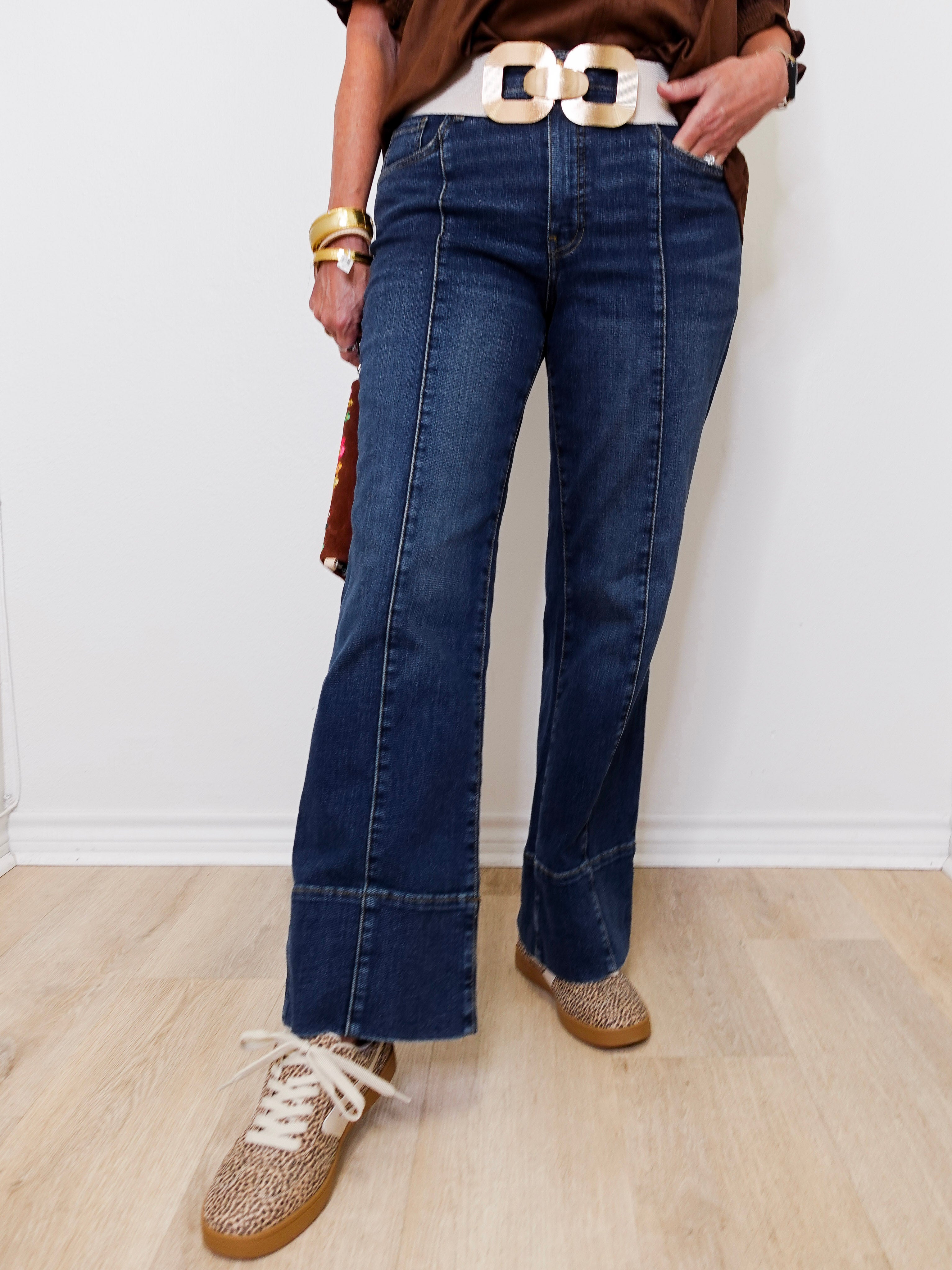 Person wearing blue jeans with a gold belt and brown shoes on a light wooden floor.
