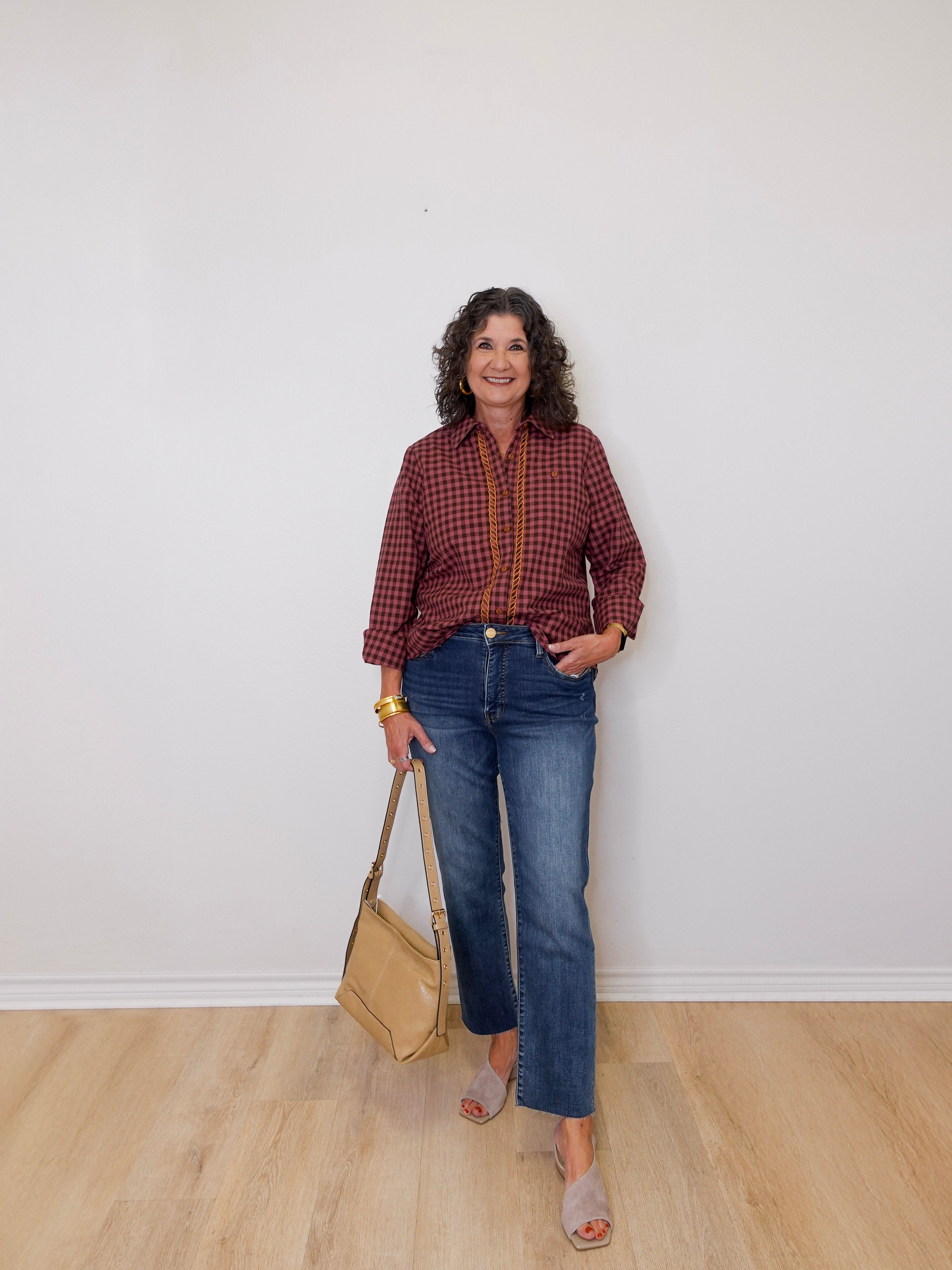 Woman wearing a plaid shirt and jeans, holding a beige handbag against a white wall.