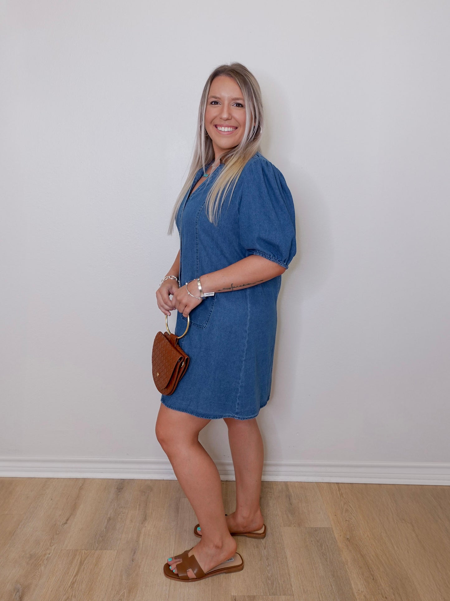 Woman wearing a blue dress holding a brown handbag against a white wall.