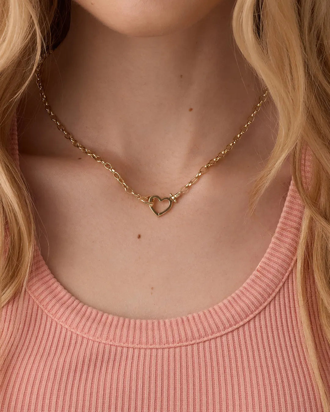 Gold heart necklace worn by a person with blonde hair and a pink top.