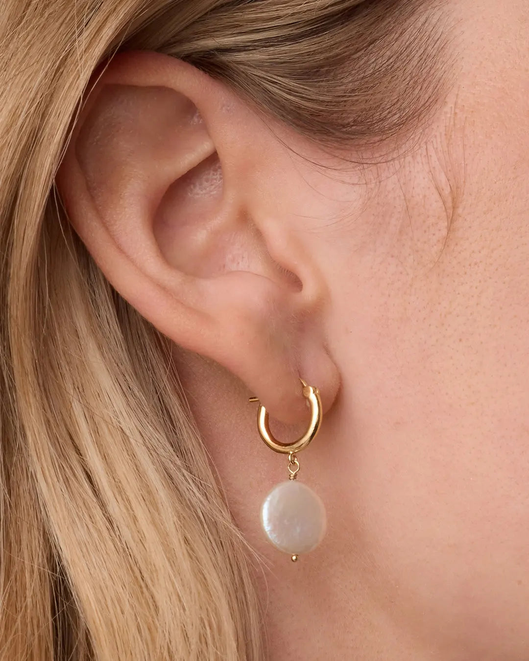 Gold hoop earring with a pearl drop worn by a person with blonde hair.