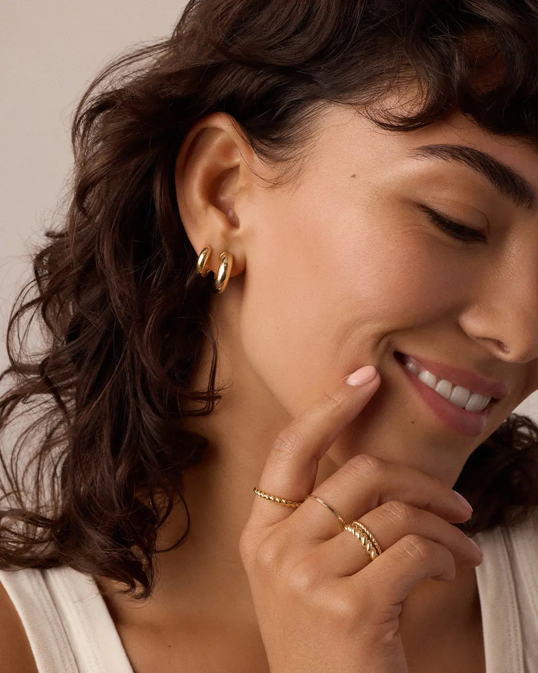 Woman wearing gold hoop earrings and rings with a neutral background