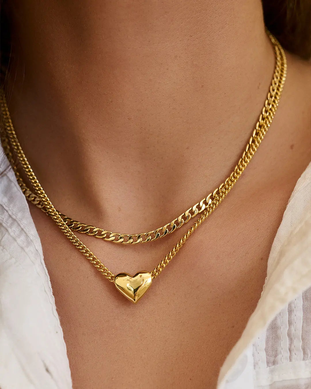 Gold chain necklace with a heart pendant on a person wearing a white top.