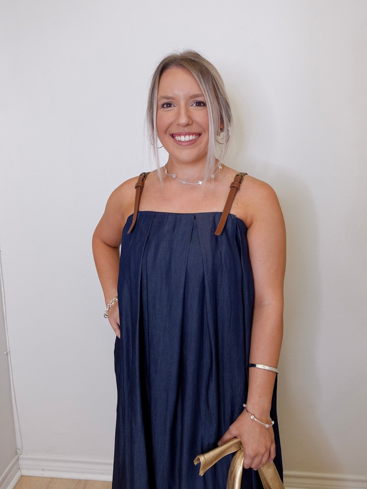 Woman wearing a navy blue dress with brown straps against a white wall.