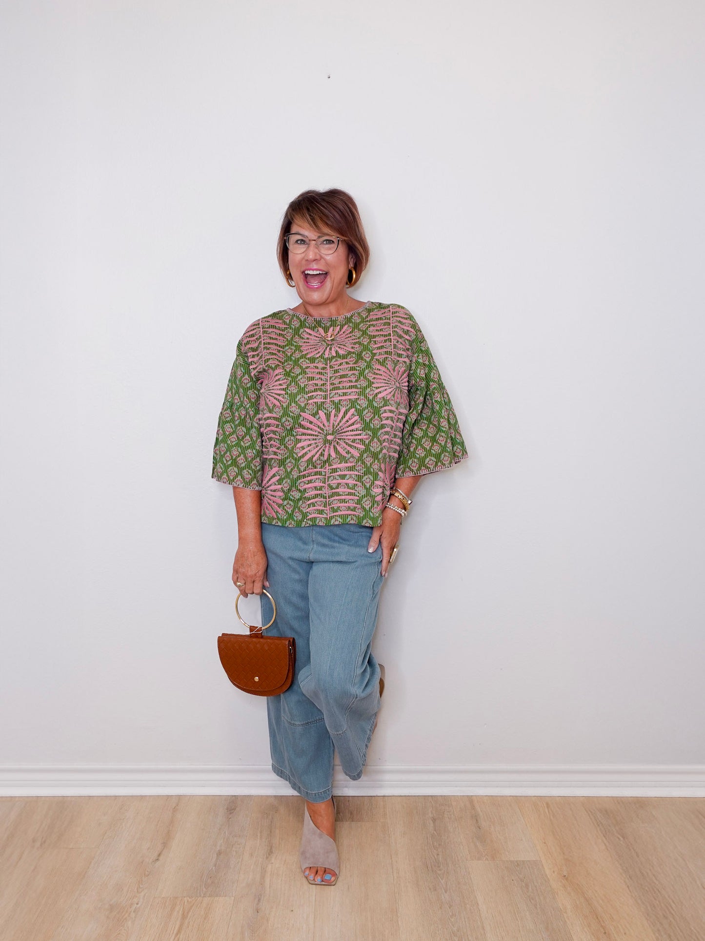 Woman wearing a patterned top and jeans, holding a handbag against a white wall.