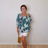 Woman wearing a patterned top and white shorts against a plain wall.