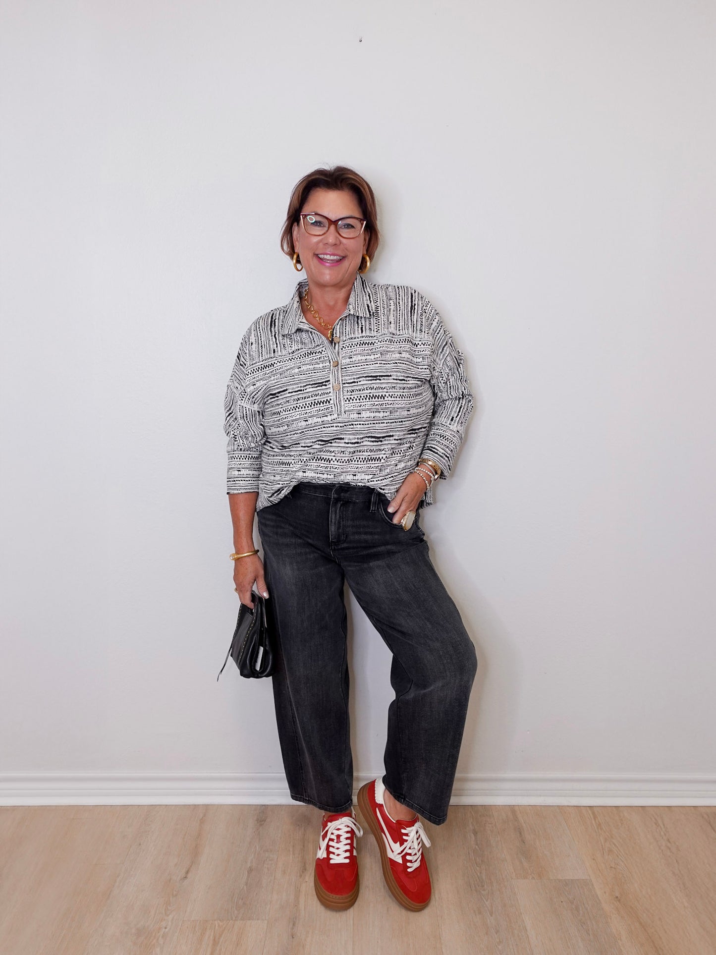 Person wearing a patterned shirt, dark jeans, and red shoes against a white wall.