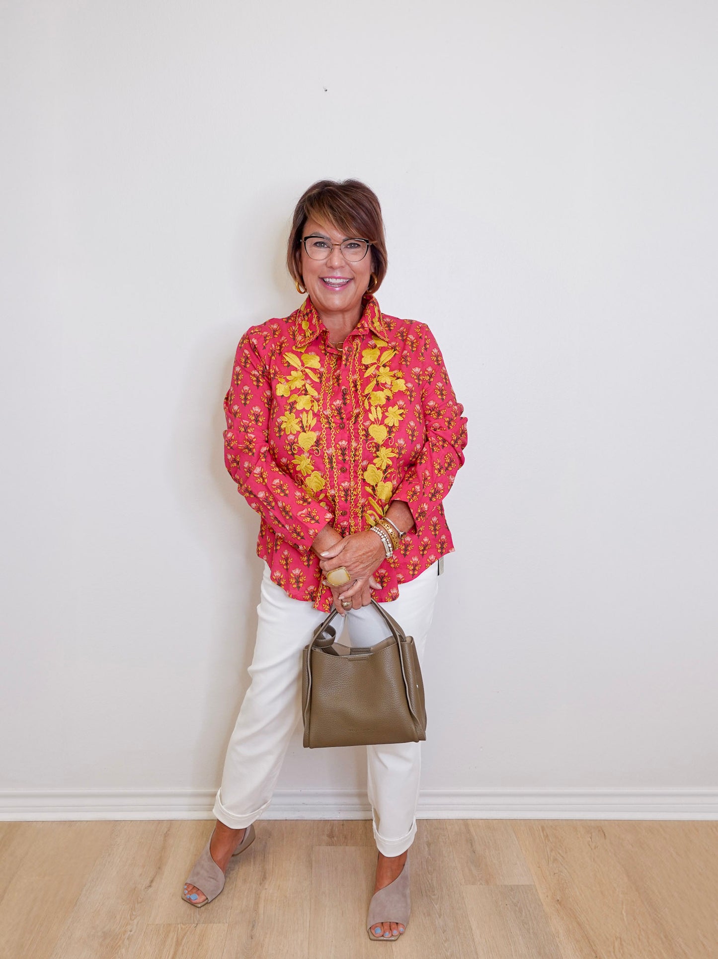 Woman wearing a pink floral blouse, white pants, and holding a handbag against a white wall.