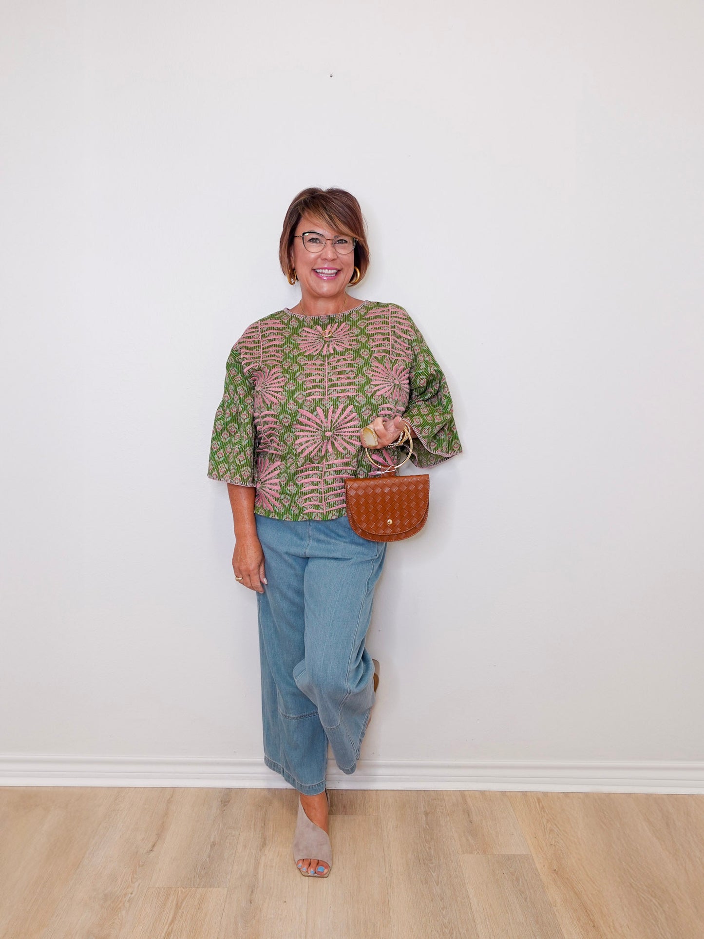 Woman wearing a green patterned top and blue jeans, holding a brown bag against a white wall.