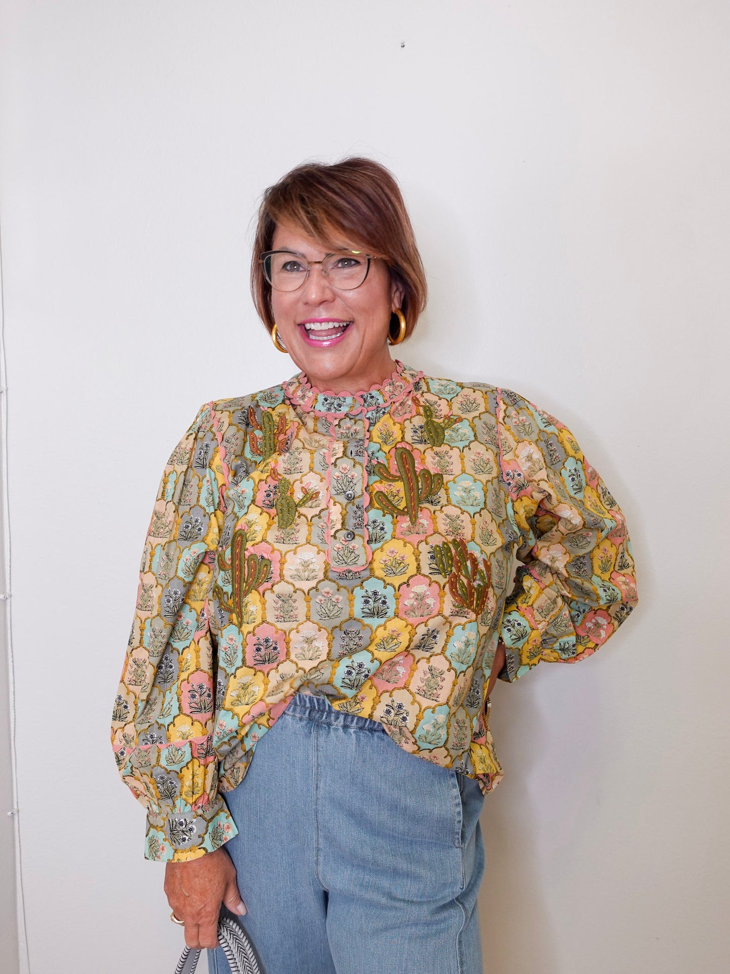 Woman wearing a patterned blouse and jeans against a white wall