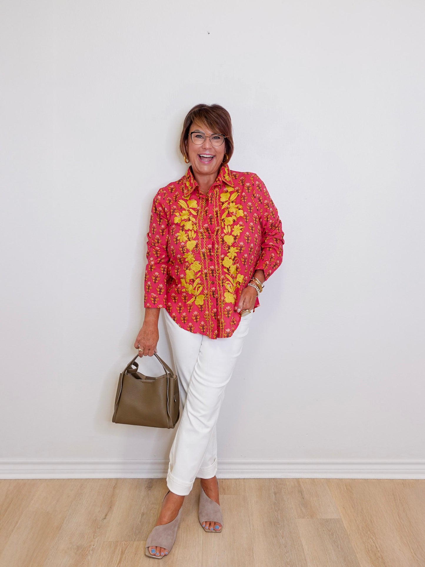 Woman wearing a pink floral blouse and white pants, standing against a plain white wall.