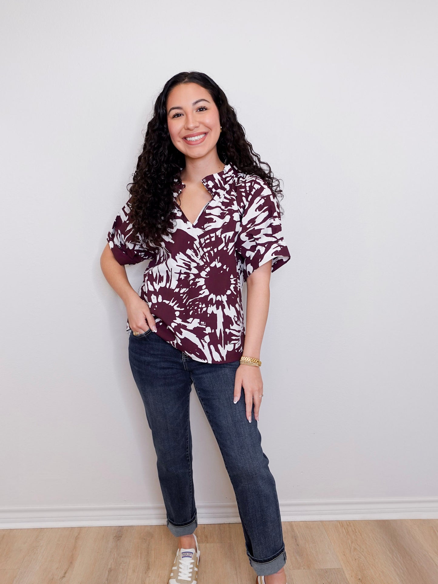 Woman wearing a patterned blouse and jeans standing against a plain wall.