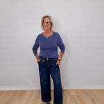 Woman wearing a purple sweater and blue jeans standing against a plain white wall.