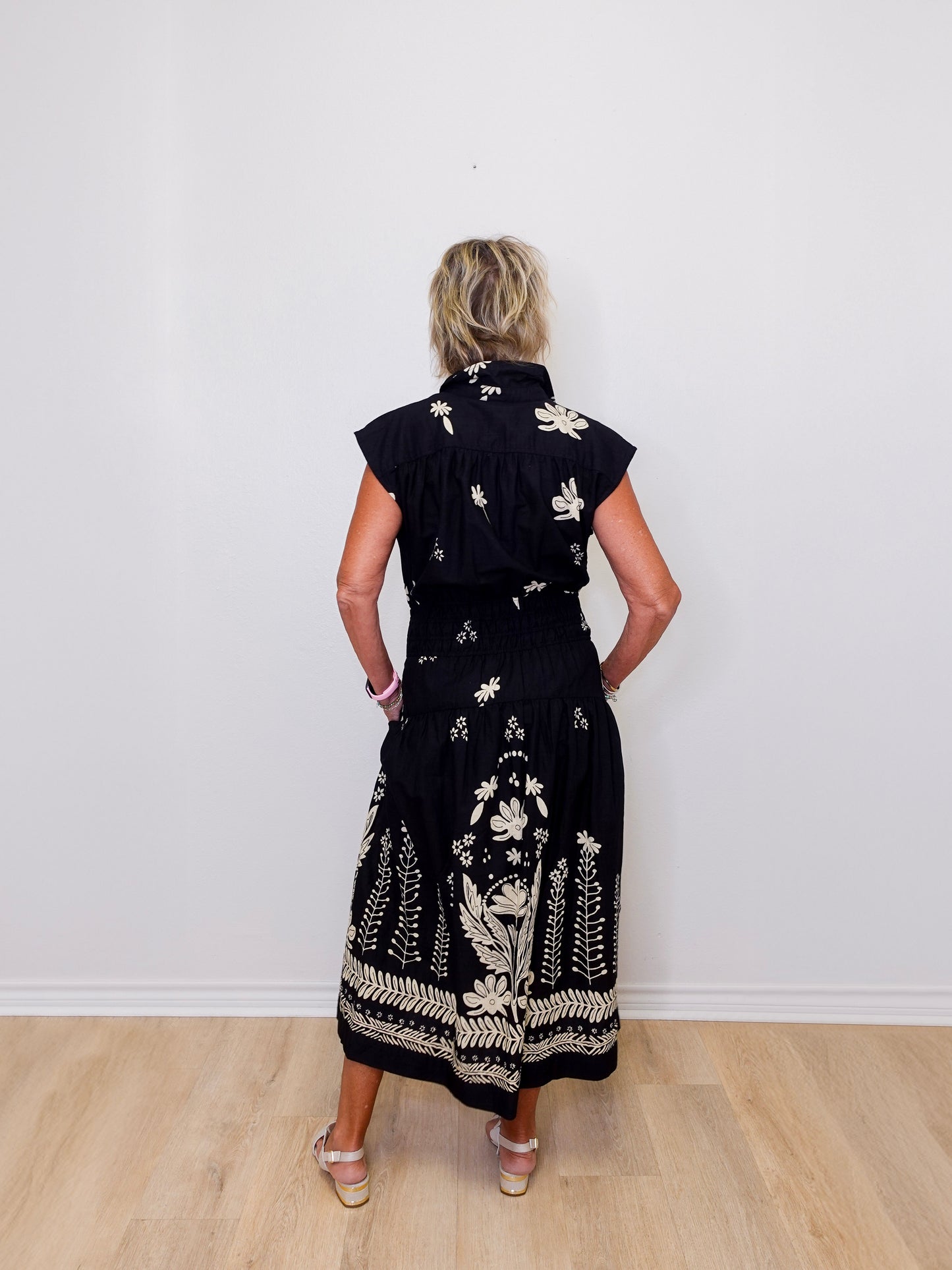 Woman wearing a black dress with white floral patterns against a white wall.