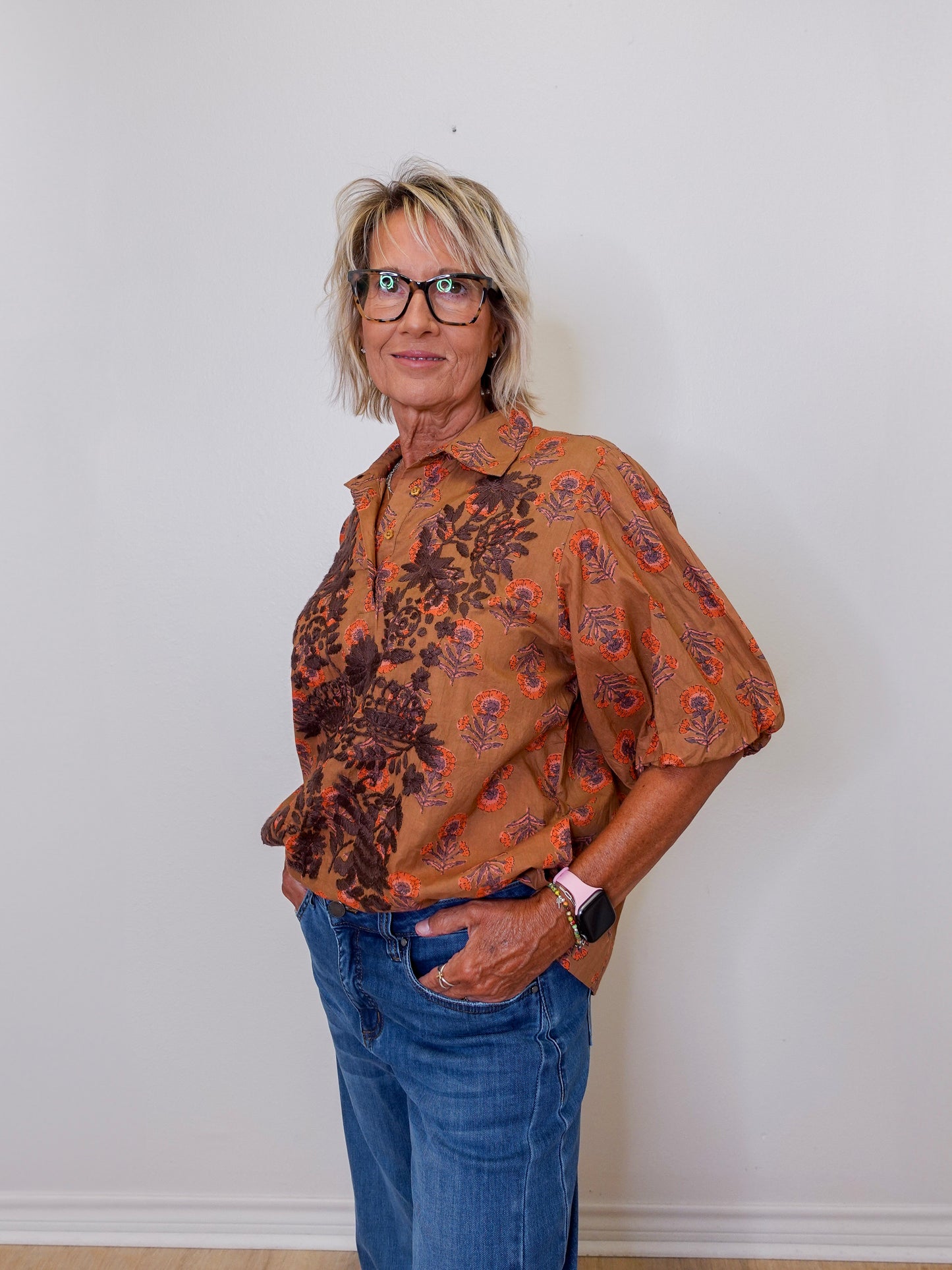 Woman wearing a patterned brown shirt and blue jeans against a white wall