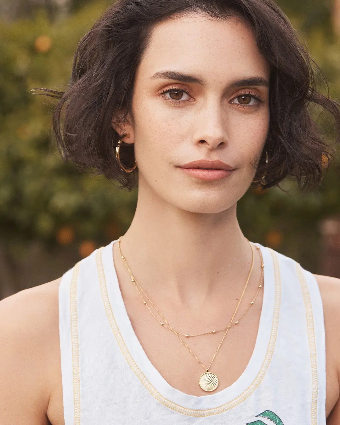 Woman wearing a white tank top with a logo, layered necklaces, and hoop earrings against a blurred natural background.