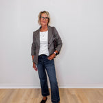 Woman standing against a white wall wearing a brown blazer, white shirt, blue jeans, and brown shoes.