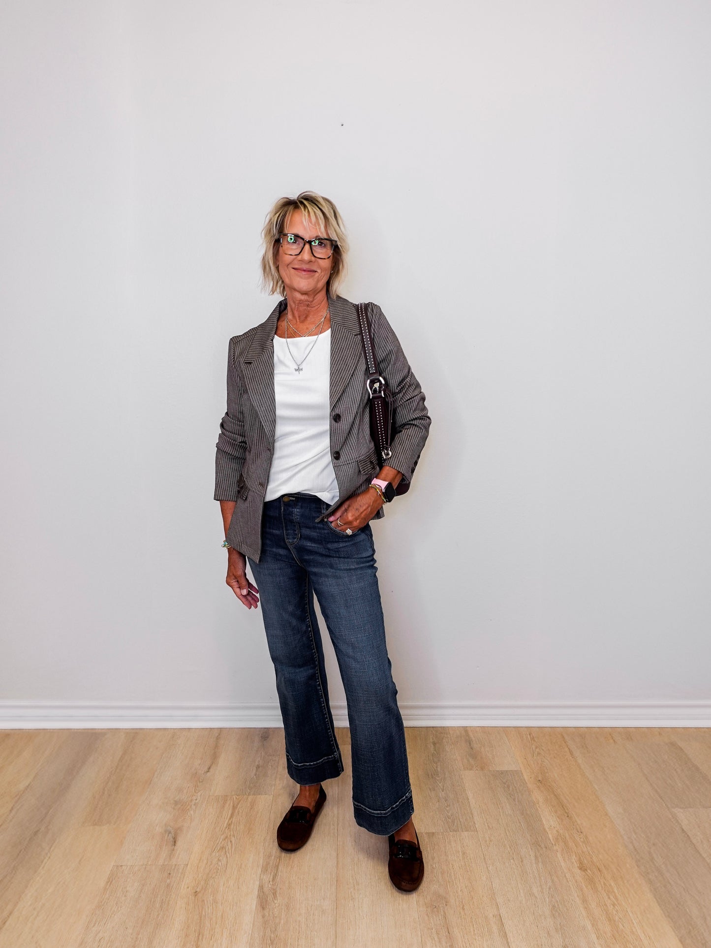 Woman standing against a white wall wearing a brown blazer, white shirt, blue jeans, and brown shoes.