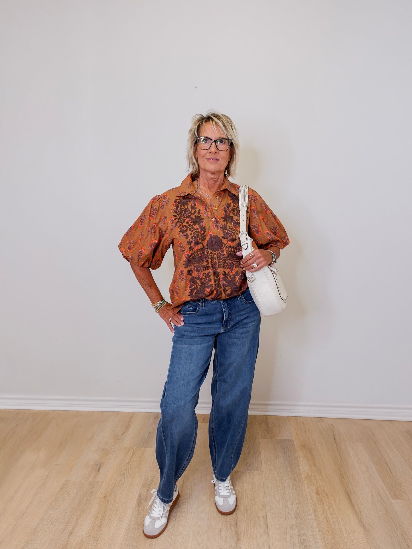 Woman wearing a patterned shirt, jeans, and white sneakers against a plain wall.