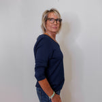 Person wearing a navy blue sweater and jeans standing against a white wall.