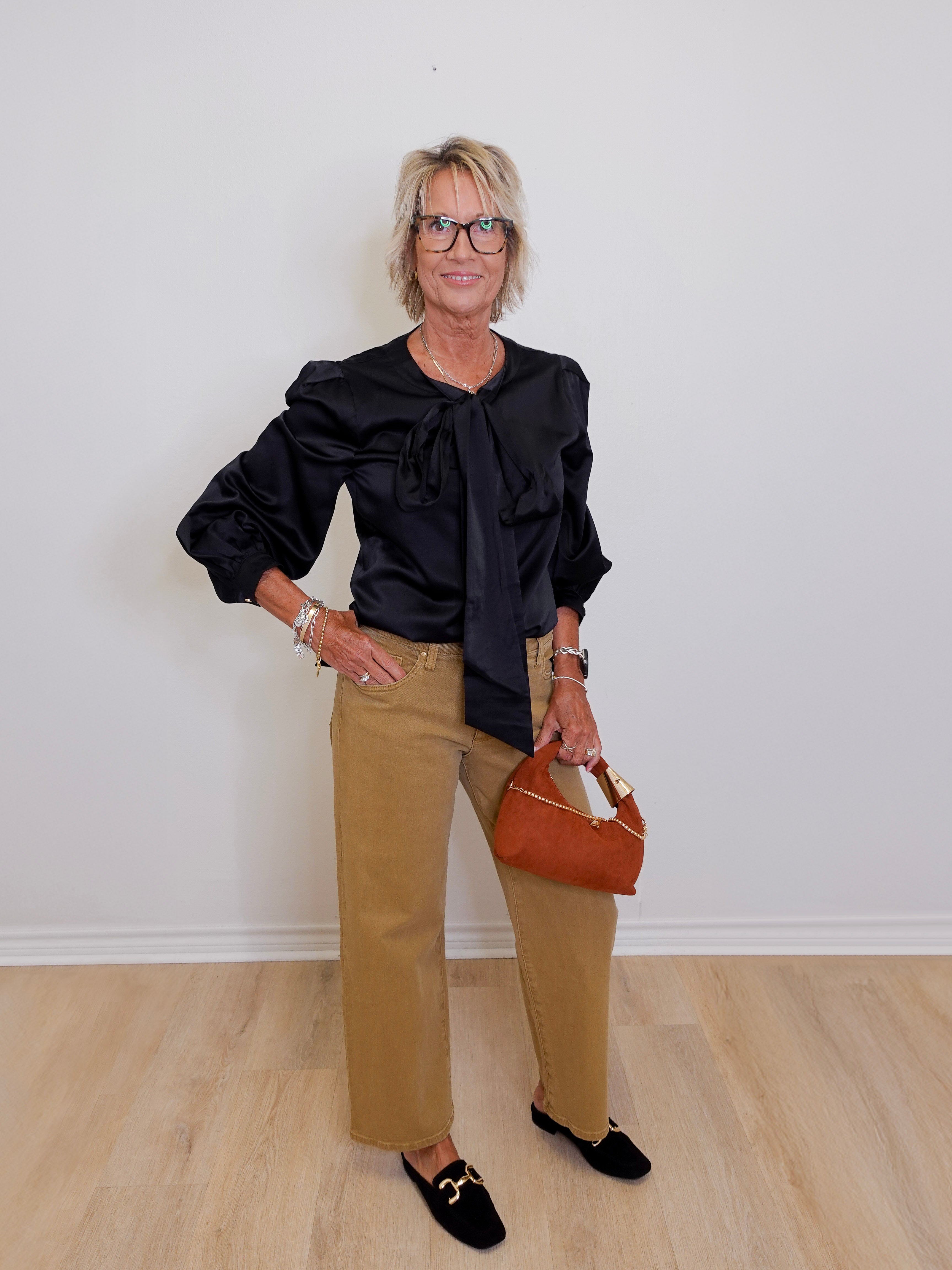 Woman wearing a black blouse with ruffled sleeves and tan pants, holding a brown handbag.