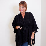 Woman wearing a black poncho and holding a patterned handbag against a white wall.