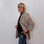 Person wearing a khaki blazer over a black top and blue jeans against a white wall.