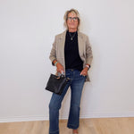 Person wearing a beige blazer, black top, blue jeans, and holding a black handbag against a white wall.