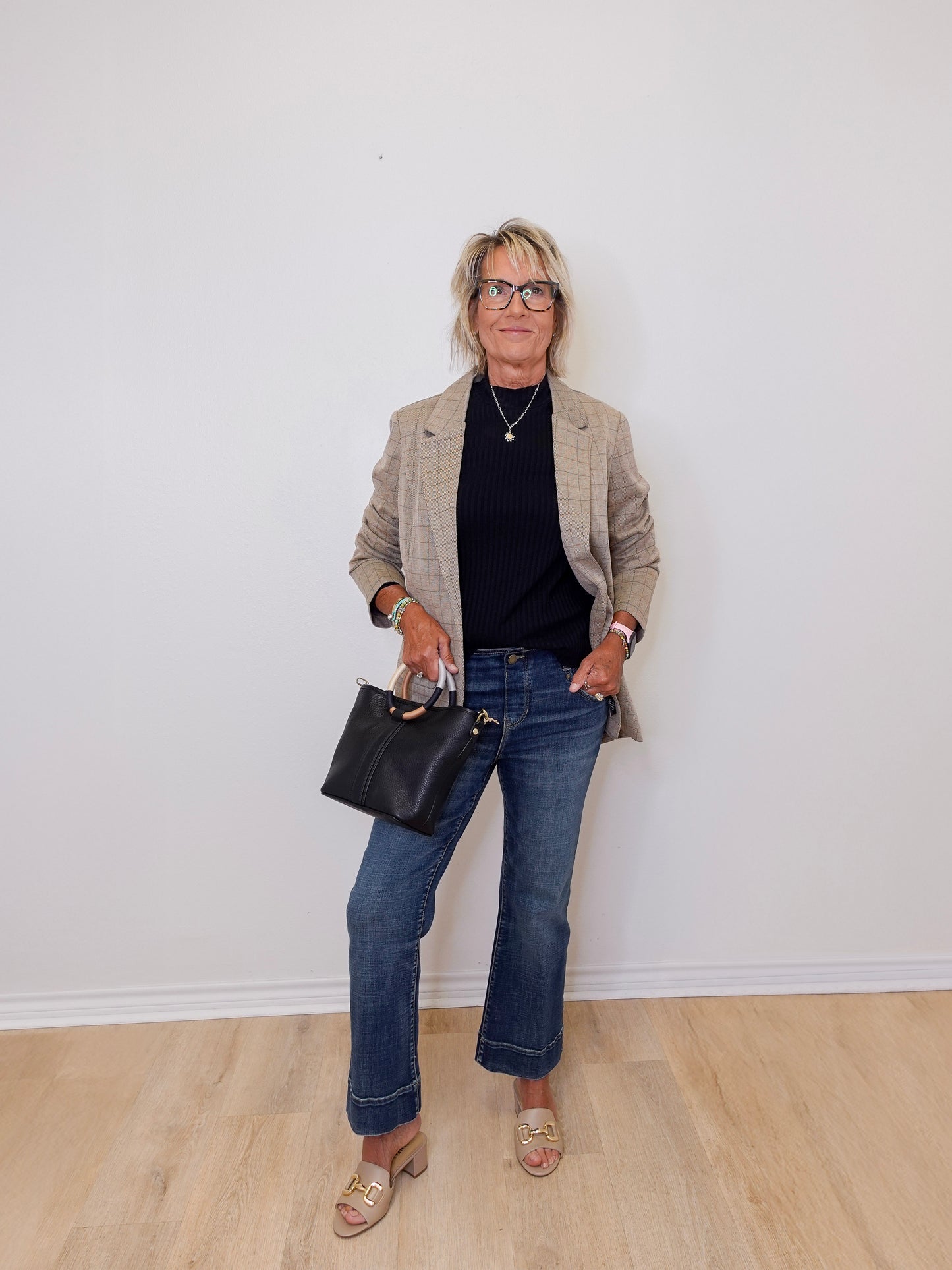 Person wearing a beige blazer, black top, blue jeans, and holding a black handbag against a white wall.