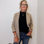 Woman wearing a plaid blazer, black top, and jeans, holding a black handbag against a white wall.