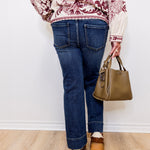 Person wearing a patterned top, blue jeans, and brown shoes, holding a green handbag against a white wall.