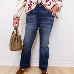 Person wearing a floral blouse, blue jeans, and brown shoes holding a beige handbag against a white wall.