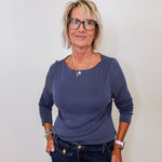 Woman wearing a blue long-sleeve top and dark jeans against a white wall