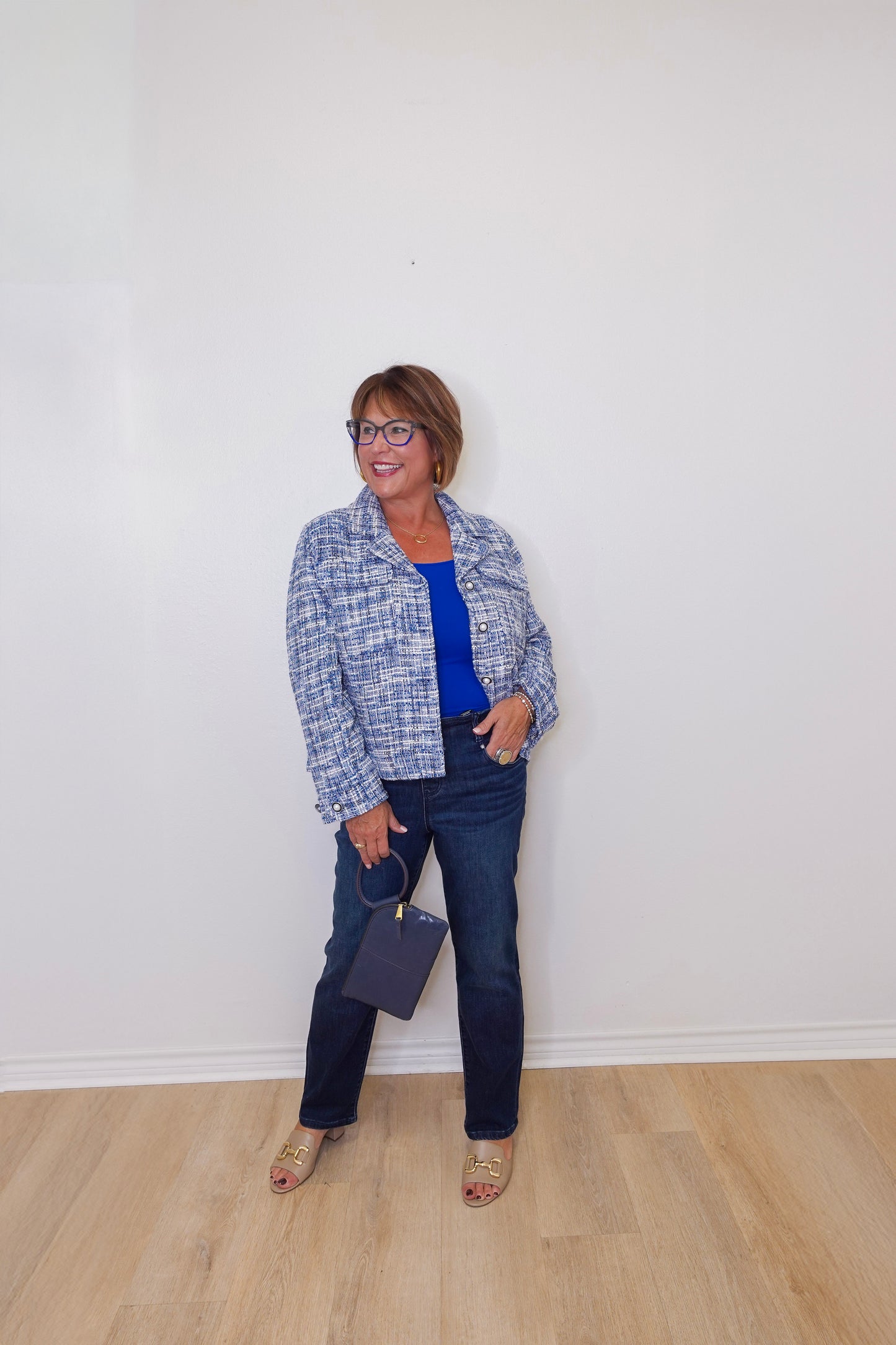 Woman standing against a white wall wearing a blue plaid jacket, blue shirt, and jeans.
