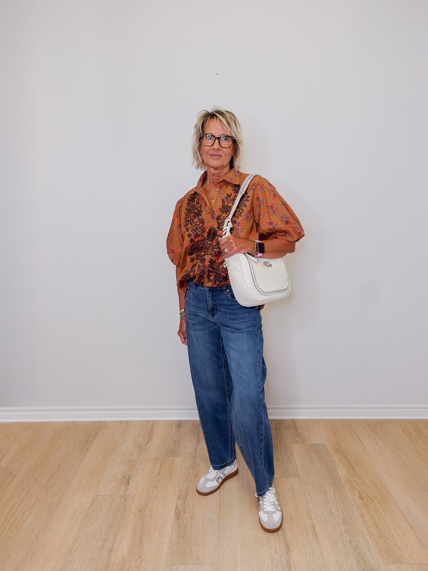 Woman wearing a patterned shirt, jeans, and white sneakers with a white handbag against a plain background.