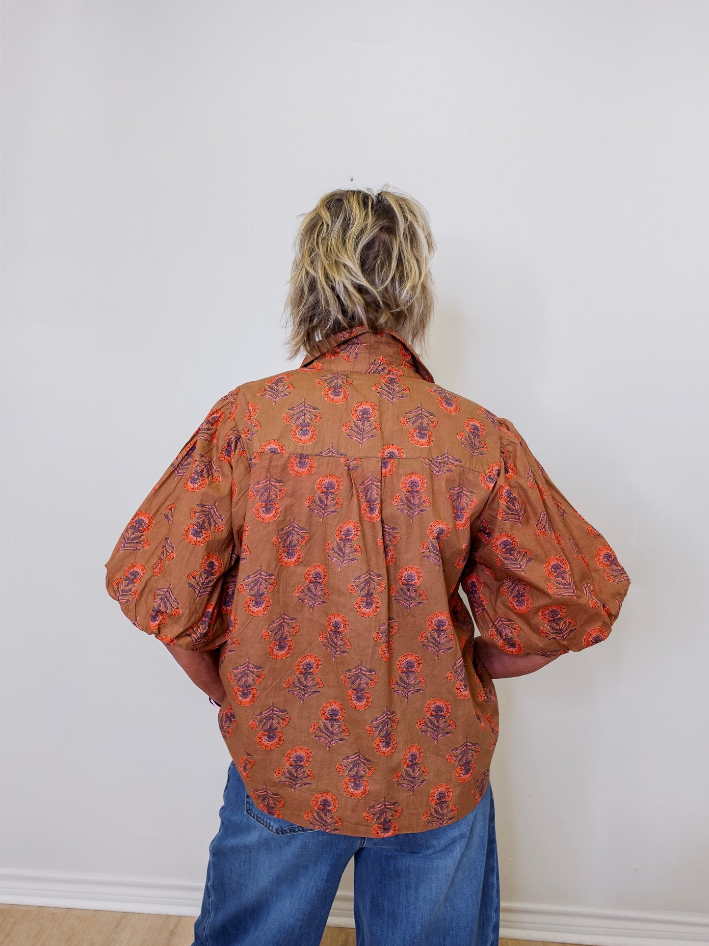 Person wearing a patterned brown shirt and blue jeans against a white wall