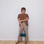 Woman standing against a white wall wearing a patterned top and beige pants, holding a blue handbag.