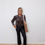 Woman wearing a leopard print top and black pants, holding a beige bag against a white wall.