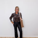 Woman wearing a leopard print top and black pants, holding a beige bag against a white wall.