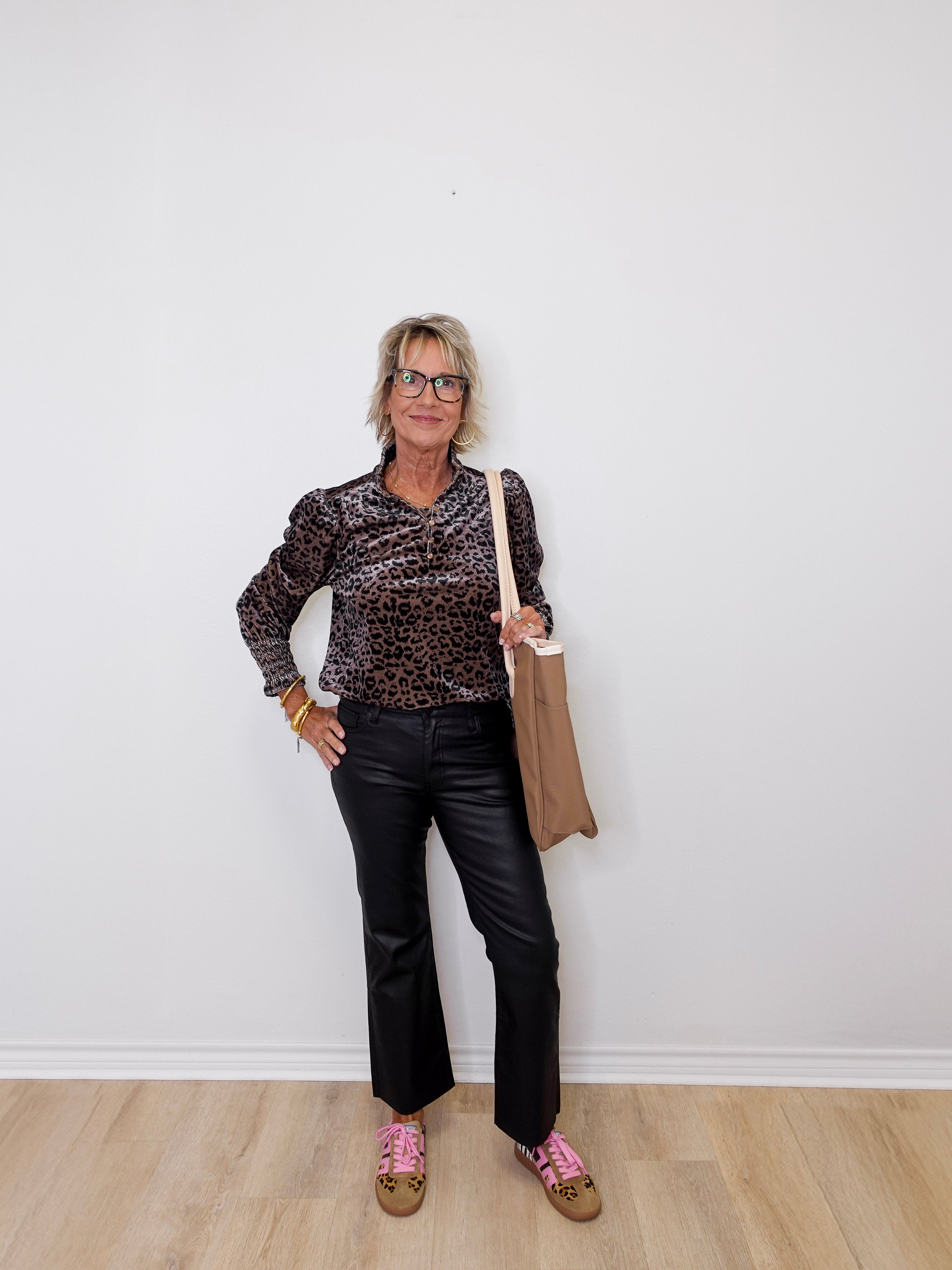 Woman wearing a leopard print top and black pants, holding a beige bag against a white wall.