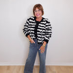 Person wearing a black and white patterned jacket, blue jeans, and black loafers against a white wall.
