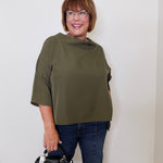 Woman wearing an olive green top and blue jeans, holding a black handbag against a white wall.