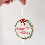 Decorative ornament with 'Baby's 1st Christmas' text and a red bow, held by a hand against a white background.