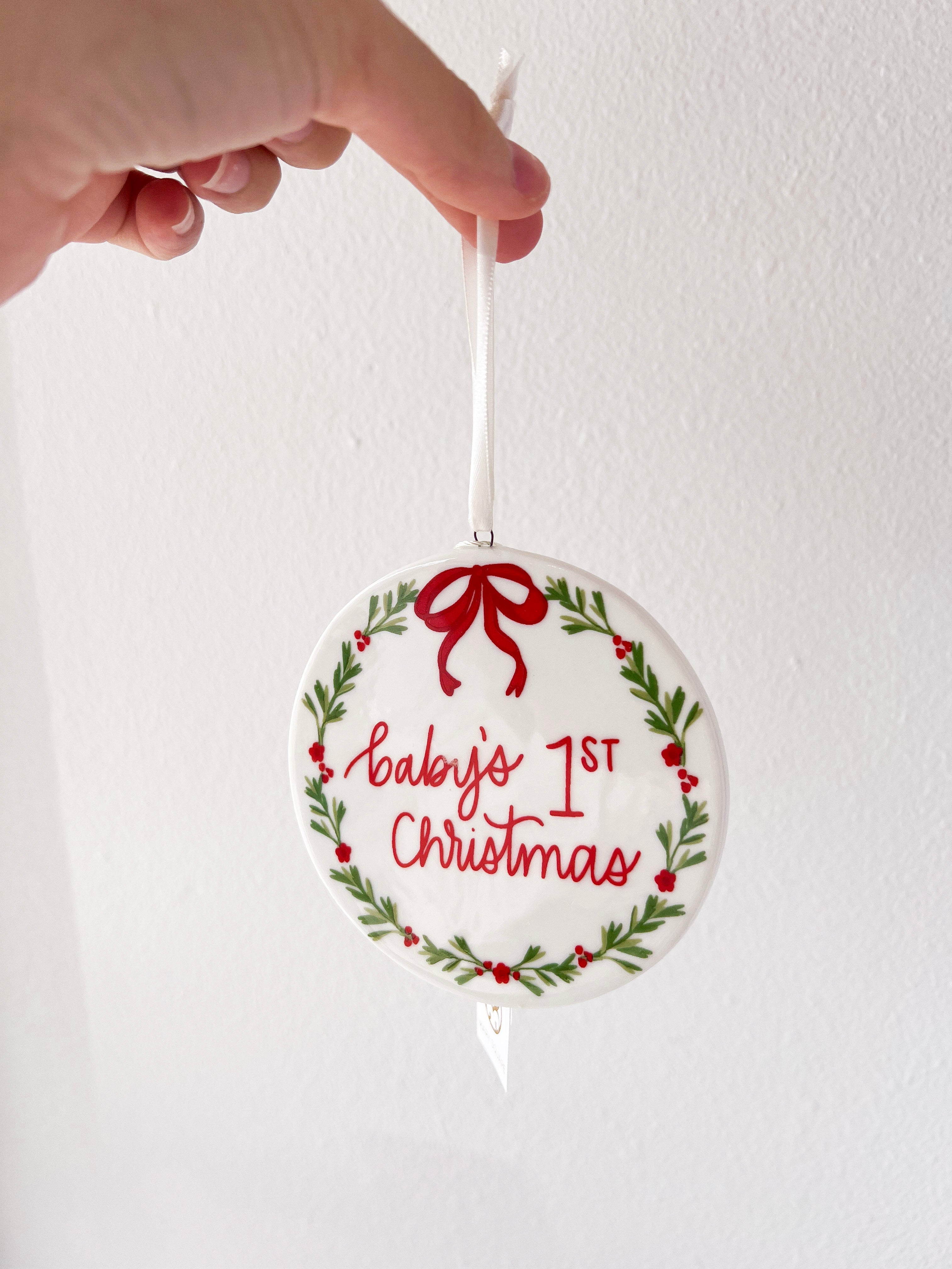 Decorative ornament with 'Baby's 1st Christmas' text and a red bow, held by a hand against a white background.