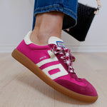 Pink Adidas sneaker with white stripes worn by a person on a light wooden floor.