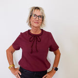 Woman wearing a burgundy blouse with a decorative bow and black pants against a white background