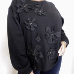 Black sweater with floral embellishments worn by a person on a white background