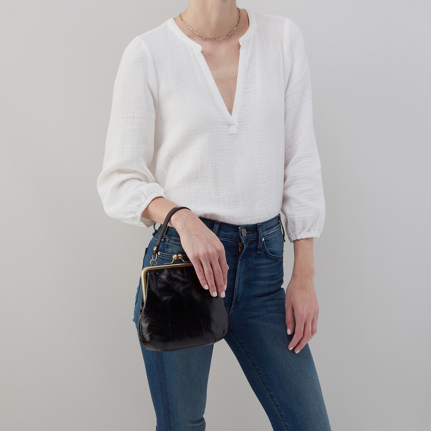 Person wearing a white sweater and blue jeans holding a black handbag against a gray background